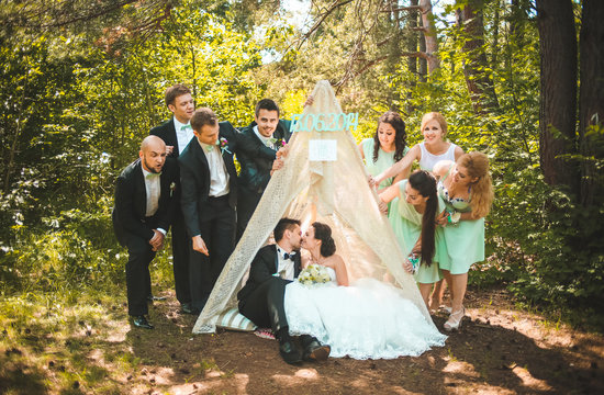 Full length portrait of newlywed couple having fun with bridesmaids and groomsmen in green sunny park. Friends near hand made tent