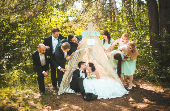 Full length portrait of newlywed couple having fun with bridesmaids and groomsmen in green sunny park. Friends near hand made tent