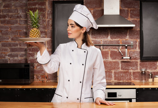 Female chef in white cap and uniform with pineapple on the plate in her hands
