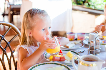 Adorable little girl having breakfast at restaurant. Cute kid enjoy fresh orange juice in outdoor cafe