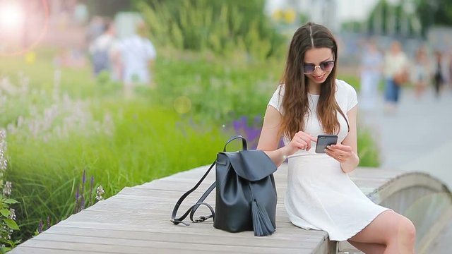Young caucasian woman sending message and listen music outdoor at european city. Beautiful girl in sunglasses sitting on wooden bench using smartphone