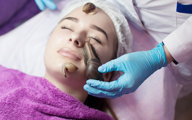 Naklejka premium Young woman receiving snail facial massage. Snail on face. Cleaning procedure in spa salon. Blurred background.