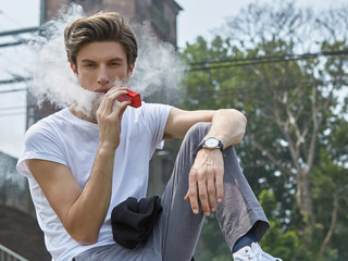Junger Mann in T-Shirt und Jeans sitzt e-Zigarette rauchend an einer Bahnstrecke und bläst lässig eine weiße Dampfwolke. - obrazy, fototapety, plakaty