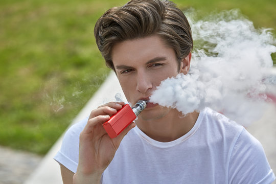 Junger Mann in T-Shirt und Jeans sitzt e-Zigarette rauchend im Sonnenschein und bl&auml;st l&auml;ssig eine wei&szlig;e Dampfwolke.
