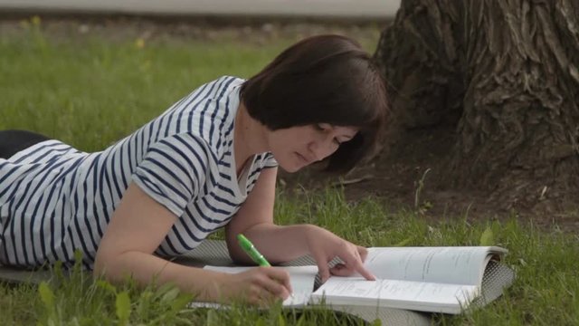 Female student doing task in the park. She styding while lying on the mat near the tree