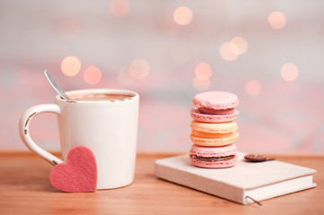 Colorful macaroons staying on book with cup of coffee and little heart over christmas lights. Good morning. Selective focus.