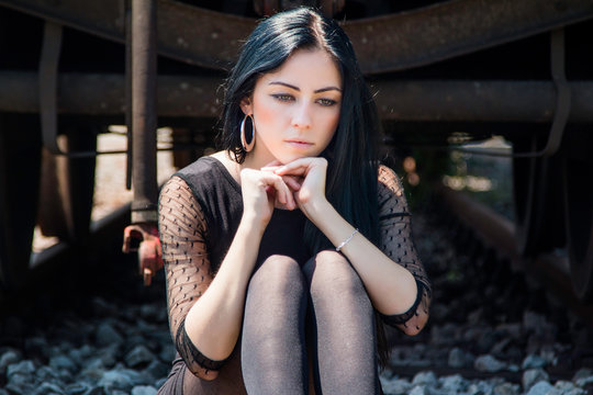      Young Beautiful Girl In Black Dress And Nylons Sitting On Rail Tracks, Cargo Wagons In Background, Emotive Portrait