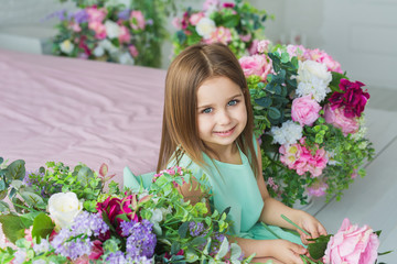 Pretty little girl in a turquoise dress sits and smiles near a flowers in a studio
