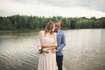 elegant stylish groom with his happy gorgeous brunette bride on the background of a lake