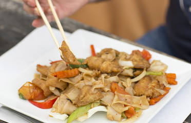 Thai food for lunch in a plate on a wooden table. Fried noodles with fish, red pepper and greens. Man eating wooden chopsticks. 