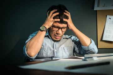Asian man feeling exhausted. Young beard man wear eyeglasses having headache while sitting at his working place in office. Stressed and depressed asian man.