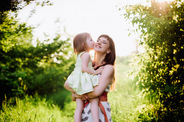 Mom kisses and hugs daughter on nature in sunset light, family, motherhood, child