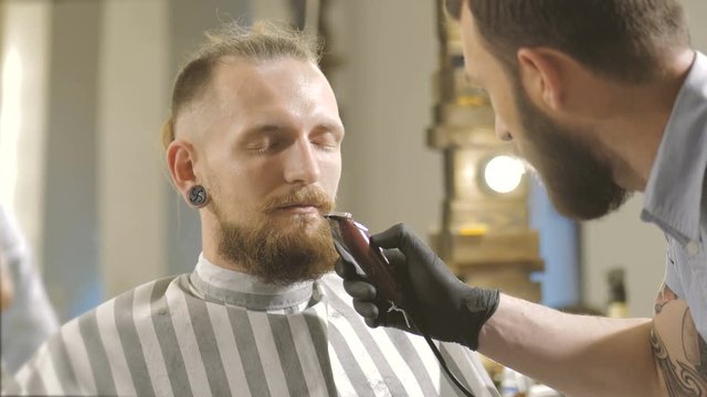Getting perfect shape. Close-up side view of young bearded man getting beard haircut by hairdresser at barbershop