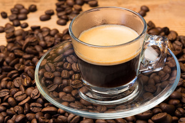A transparent glass cup of hot espresso on a saucer standing on a wooden table top, surrounded by coffee beans