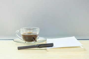 Closeup black pen on white book with black coffee in transparent cup of coffee on blurred wooden desk and frosted glass wall textured background