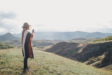 Young beautiful woman traveler wearing hat and poncho taking pictures on her smart phone standing on the top of the mountain