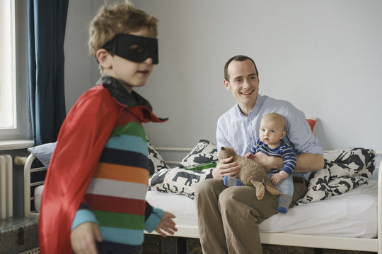 Boy wearing cape standing in front of father and brother at home