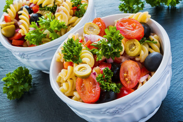 Salad: pasta fusilli, black and green olives, cherry tomatoes, red onion and parsley. Dressing: olive oil and lemon juice. In white bowl. Black stone background.