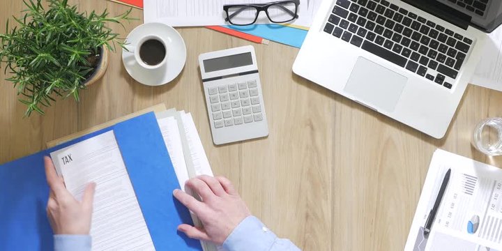 Businessman working at office desk, he is checking tax forms and using a calculator, top view