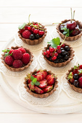 Close up of chocolate tartlets with chocolate cream, fresh strawberries, raspberries, blueberries, red currants and cherries on white wooden background. Selective focus