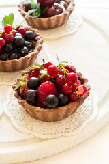Close up of chocolate tartlets with chocolate cream, fresh strawberries, raspberries, blueberries, red currants and cherries on white wooden background. Selective focus