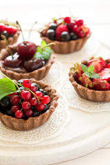 Close up of chocolate tartlets with chocolate cream, fresh strawberries, raspberries, blueberries, red currants and cherries on white wooden background. Selective focus