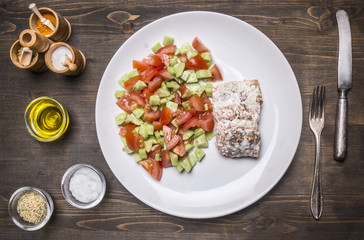 fried red fish with sauce and garnished with vegetable salad on white plate with spices and vintage knife and fork on wooden rustic background top view