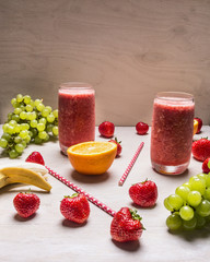 Red smoothie drink in glass with straw and fruits berries ingredients on white wooden background, top view with strawberries, white grapes, bananas