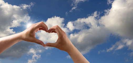 Liebe Symbol Hände zum Herz geformt vor blauem Himmel mit Wolken und Sonnenschein