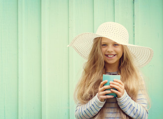 Cute little girl stands near a turquoise wall in white hat and holding cup. Space for text