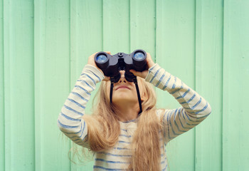 Cute little girl stands near a turquoise wall and looks binoculars. Space for text