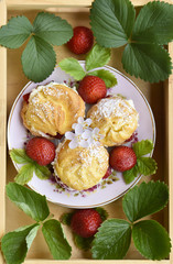 Homemade cream puffs or profiterole filled with whipped cream, powdered sugar topping served with strawberries in a wooden tray