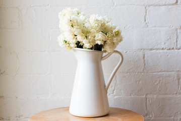 Erlicheer daffodils in white jug on wooden table centred against rustic white brick wall
