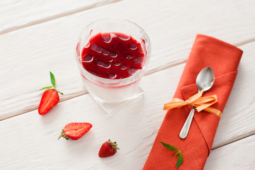 Strawberry organic panna cotta in glass with spoon flat lay. Dessert panna cotta with fresh strawberry slices served in glass and spoon on orange napkin on white wooden table