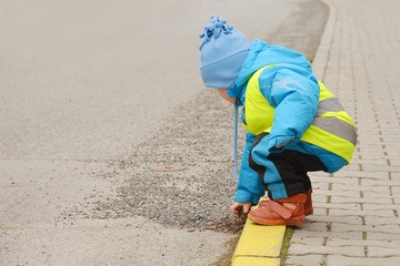 Small child playing with a stones on a roadside alone. He is marked by reflective vest and the...