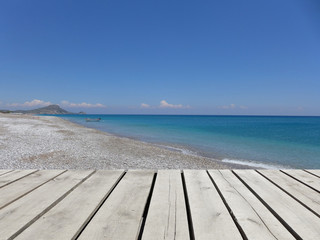Wooden pier with blue sea, sky and white pebble beach background. Mediterranean landscape in sunny day. Greek island.