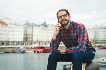 Young bearded man talking on phone while sitting at leissure harbor. Urban maritime vacations lifestyle.