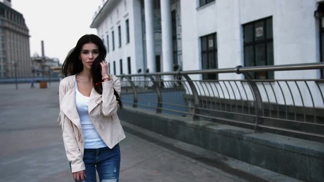 Beautiful woman with long dark hair walking on city street wearing jeans and white jacket