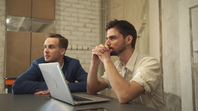 Two men sitting at the desk with computer waiting client. Partners in creative office. Young caucasian man wearing in blue jackets. Mixed race guy dressed in casual shirt rolled up sleeves. Young