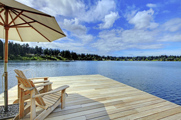Two adirondack wooden chairs with umbrella on dock facing blue lake.