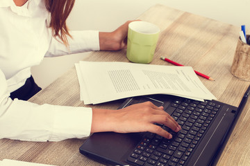 Mujer trabajando con un ordenador escribiendo y con papeles y una taza de café sobre un escritorio de madera. Vista superior y de cerca - obrazy, fototapety, plakaty