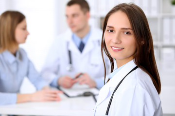 Young beautiful female doctor smiling  on the background with patient and his doctor in hospital. High level and quality medical service concept.