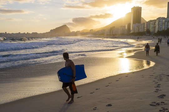 Silhouette of bodyboarder walking in front of golden sun setting behind the Rio de Janeiro skyline at the Leme end of Copacabana Beach