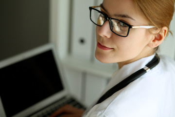 Young brunette female doctor standing with laptop computer  near window in  hospital.  Physician ready to examine patient