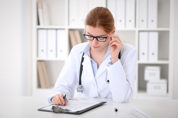 Fototapeta na wymiar Young brunette female doctor sitting at the table and working at hospital office. Health care, insurance and help concept. Physician ready to examine patient