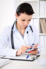 Young brunette female doctor sitting  with clipboard near window in  hospital and filling up medical history form.