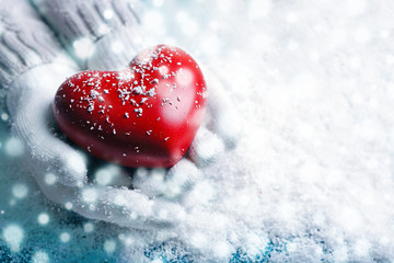 Hands in warm white gloves holding red heart on snowy background. Snow effect