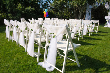 White decorated chairs on a green lawn. Chairs set in rows for the wedding ceremony. They are...