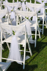 White chair with a small flower. White decorated chairs on a green lawn. Chairs set in rows for the wedding ceremony. They are decorated for the festive event. Chairs are on the green lawn outside.