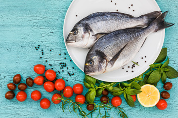 Two fresh dorado fish on white plate and vegetables on blue table. Top view, copy space. 